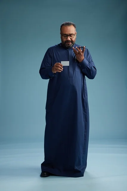 بورتريه لرجل سعودي يرتدي ثوباً أزرق ينظر إلى بطاقة عمل ممسكاً بالهاتف المحمول, الجسم بالطول الكامل, خلفيه زرقاء