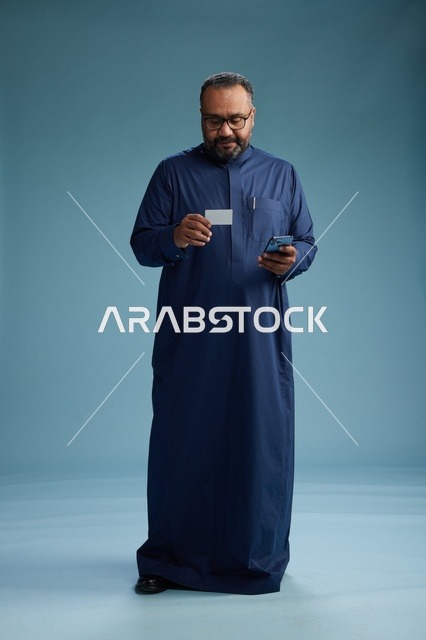 بورتريه لرجل سعودي يرتدي ثوباً أزرق ينظر إلى بطاقة عمل ممسكاً بالهاتف المحمول, الجسم بالطول الكامل, خلفيه زرقاء