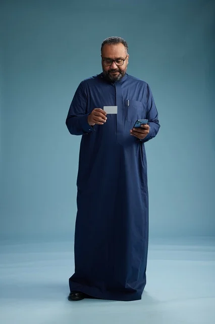 بورتريه لرجل سعودي يرتدي ثوباً أزرق ينظر إلى بطاقة عمل ممسكاً بالهاتف المحمول, الجسم بالطول الكامل, خلفيه زرقاء