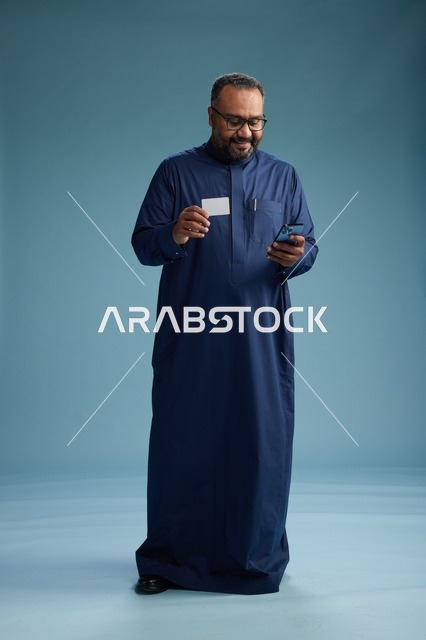 بورتريه لرجل سعودي يرتدي ثوباً أزرق ينظر إلى بطاقة عمل ممسكاً بالهاتف المحمول, الجسم بالطول الكامل, خلفيه زرقاء