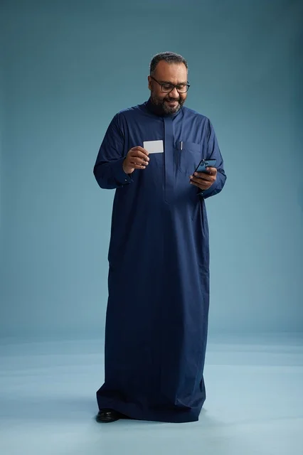 بورتريه لرجل سعودي يرتدي ثوباً أزرق ينظر إلى بطاقة عمل ممسكاً بالهاتف المحمول, الجسم بالطول الكامل, خلفيه زرقاء