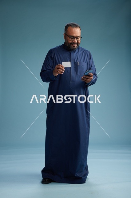 بورتريه لرجل سعودي يرتدي ثوباً أزرق ينظر إلى بطاقة عمل ممسكاً بالهاتف المحمول, الجسم بالطول الكامل, خلفيه زرقاء