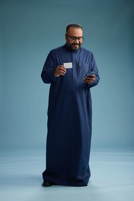 بورتريه لرجل سعودي يرتدي ثوباً أزرق ينظر إلى بطاقة عمل ممسكاً بالهاتف المحمول, الجسم بالطول الكامل, خلفيه زرقاء