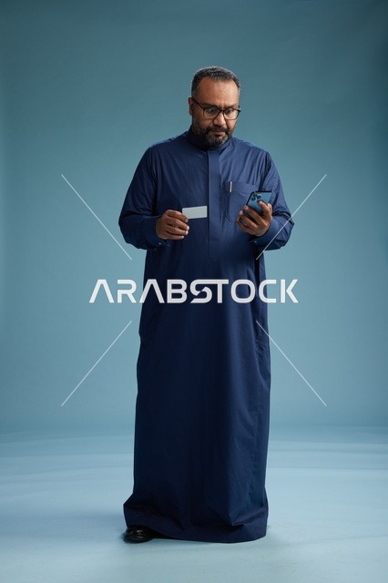 بورتريه لرجل سعودي يرتدي ثوباً أزرق ينظر إلى بطاقة عمل ممسكاً بالهاتف المحمول, الجسم بالطول الكامل, خلفيه زرقاء