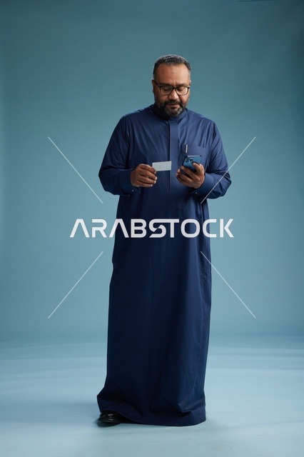 بورتريه لرجل سعودي يرتدي ثوباً أزرق ينظر إلى بطاقة عمل ممسكاً بالهاتف المحمول, الجسم بالطول الكامل, خلفيه زرقاء