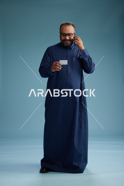 A portrait of a Saudi man wearing a blue thobe looking at a business card while holding a mobile phone, full body, blue background.
