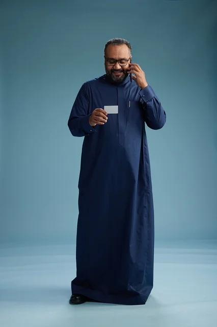 بورتريه لرجل سعودي يرتدي ثوباً أزرق ينظر إلى بطاقة عمل ممسكاً بالهاتف المحمول, الجسم بالطول الكامل, خلفيه زرقاء