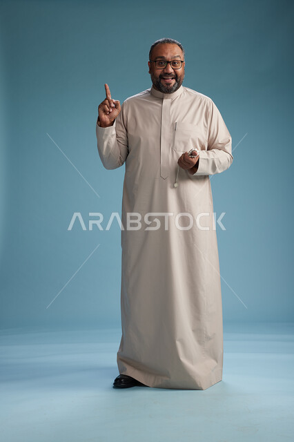 بورتريه لرجل عربي سعودي خليجي مبتسم يرتدي ثوب بيج يمسك بيده سبحه وينظر الى الكاميرا, كامل الجسم, خلفيه زرقاء
