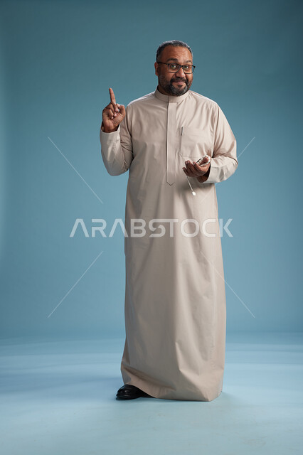 بورتريه لرجل عربي سعودي خليجي مبتسم يرتدي ثوب بيج يمسك بيده سبحه وينظر الى الكاميرا, كامل الجسم, خلفيه زرقاء
