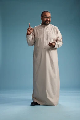 A portrait of a smiling Arab Saudi Gulf man wearing a beige thobe, holding a rosary in his hand and looking at the camera, full body, with a blue background.