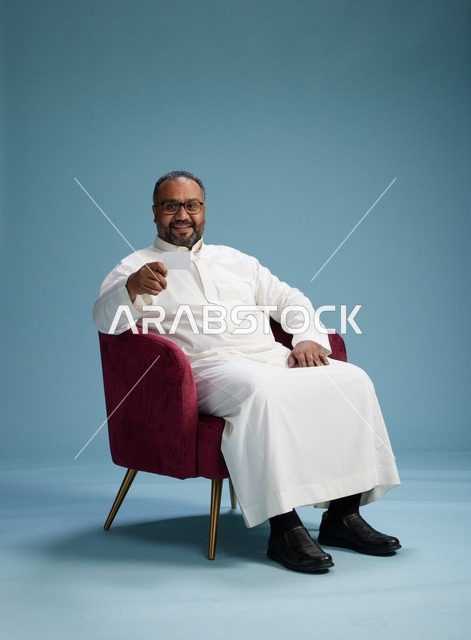 بورتريه لرجل  عربي سعودي خليجي يرتدي ثوب ابيض يحمل بطاقة بيضاء فارغه يجلس على كرسي احمر, خلفيه زرقاء