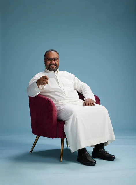 A portrait of an Arab Saudi Gulf man wearing a white thobe holding a blank white card sitting on a red chair, with a blue background.