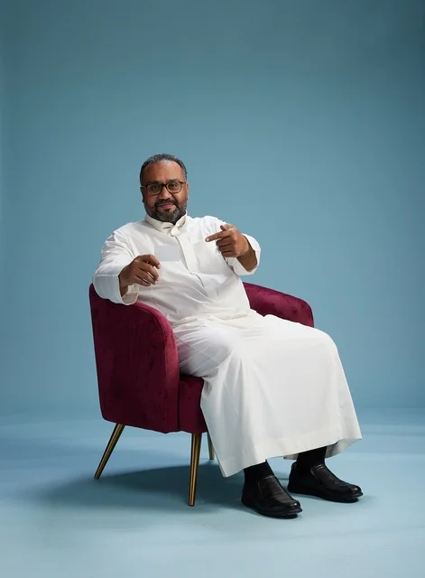 A portrait of an Arab Saudi Gulf man wearing a white thobe holding a blank white card sitting on a red chair, with a blue background.