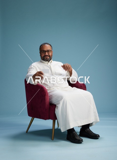 بورتريه لرجل  عربي سعودي خليجي يرتدي ثوب ابيض يحمل بطاقة بيضاء فارغه يجلس على كرسي احمر, خلفيه زرقاء