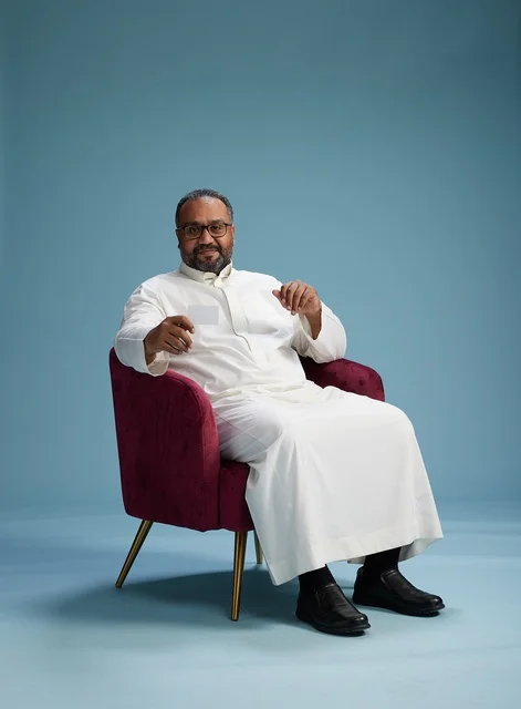 A portrait of an Arab Saudi Gulf man wearing a white thobe holding a blank white card sitting on a red chair, with a blue background.