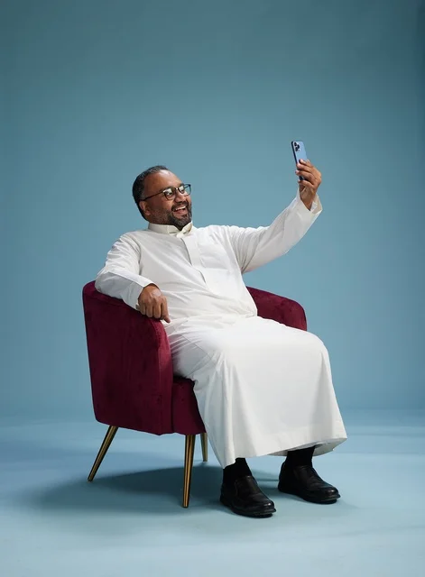 A smiling Saudi man wearing a white thobe during a video call on a mobile phone, selfie shot, blue background.