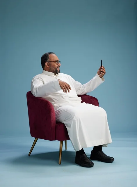 A smiling Saudi man wearing a white thobe during a video call on a mobile phone, selfie shot, blue background.