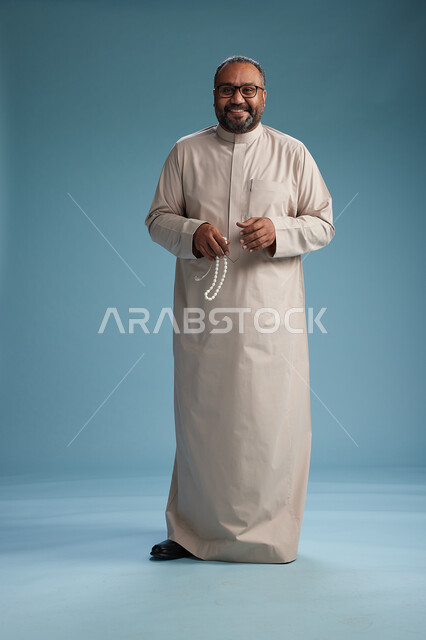 بورتريه لرجل عربي سعودي خليجي مبتسم يرتدي ثوب بيج يمسك بيده سبحه وينظر الى الكاميرا, كامل الجسم, خلفيه زرقاء