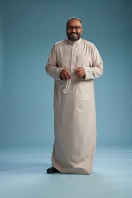 بورتريه لرجل عربي سعودي خليجي مبتسم يرتدي ثوب بيج يمسك بيده سبحه وينظر الى الكاميرا, كامل الجسم, خلفيه زرقاء