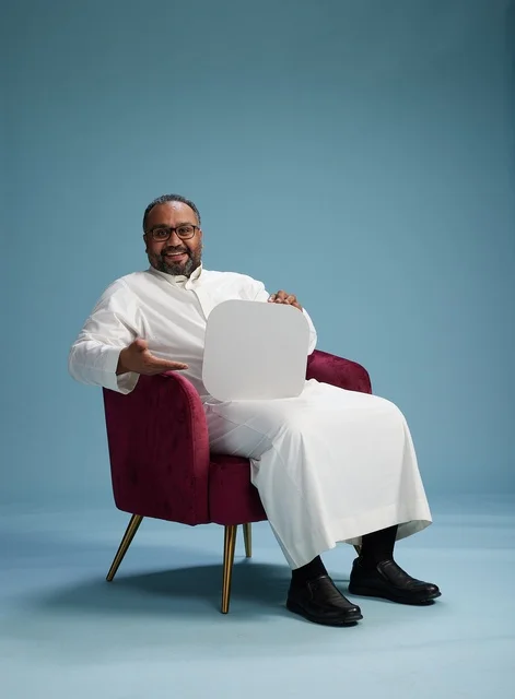 A Saudi man is holding a blank sign while sitting on a red chair, with a blank white square and a blue background.