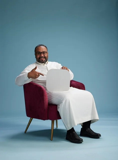 A Saudi man is holding a blank sign while sitting on a red chair, with a blank white square and a blue background.