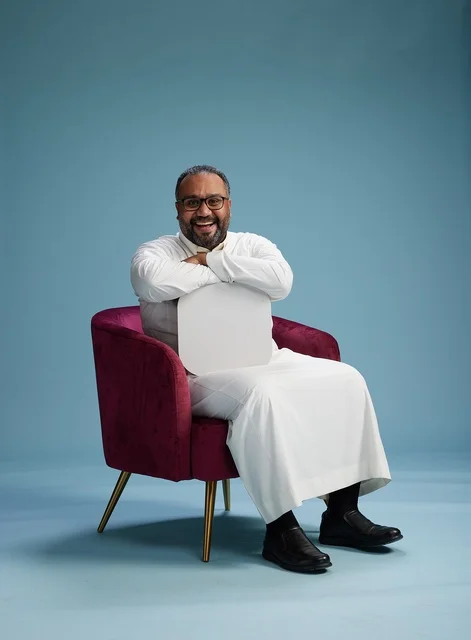 A Saudi man is holding a blank sign while sitting on a red chair, with a blank white square and a blue background.