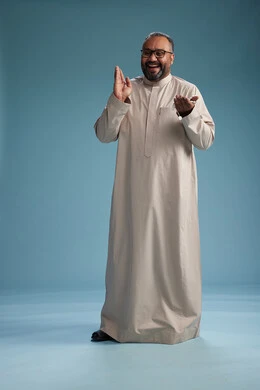 A portrait of a smiling Arab Saudi Gulf man wearing a beige thobe, holding a rosary in his hand and looking at the camera, full body, with a blue background.