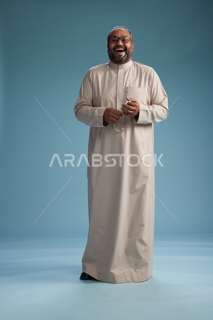 بورتريه لرجل عربي سعودي خليجي مبتسم يرتدي ثوب بيج يمسك بيده سبحه وينظر الى الكاميرا, كامل الجسم, خلفيه زرقاء
