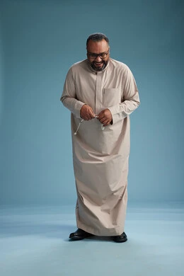 A portrait of a smiling Arab Saudi Gulf man wearing a beige thobe, holding a rosary in his hand and looking at the camera, full body, with a blue background.