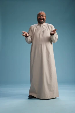 A portrait of a smiling Arab Saudi Gulf man wearing a beige thobe, holding a rosary in his hand and looking at the camera, full body, with a blue background.
