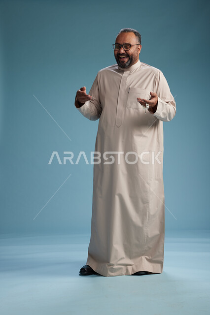 بورتريه لرجل عربي سعودي خليجي مبتسم يرتدي ثوب بيج يمسك بيده سبحه وينظر الى الكاميرا, كامل الجسم, خلفيه زرقاء