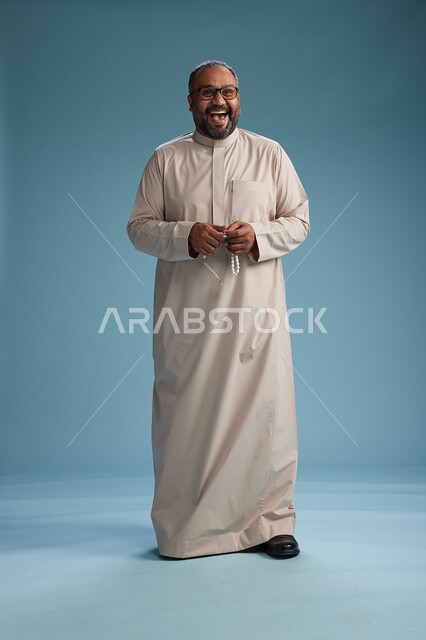 بورتريه لرجل عربي سعودي خليجي مبتسم يرتدي ثوب بيج يمسك بيده سبحه وينظر الى الكاميرا, كامل الجسم, خلفيه زرقاء