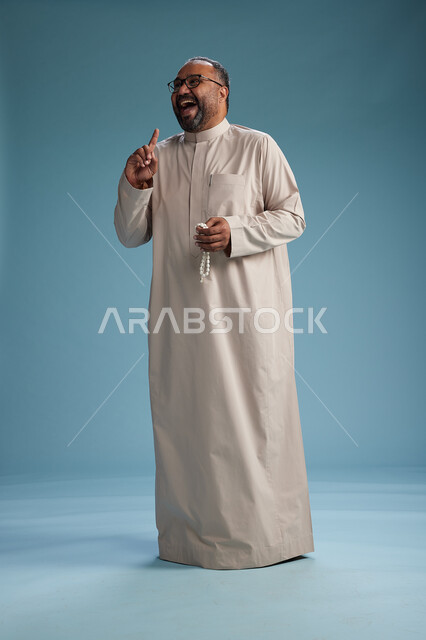 بورتريه لرجل عربي سعودي خليجي مبتسم يرتدي ثوب بيج يمسك بيده سبحه وينظر الى الكاميرا, كامل الجسم, خلفيه زرقاء