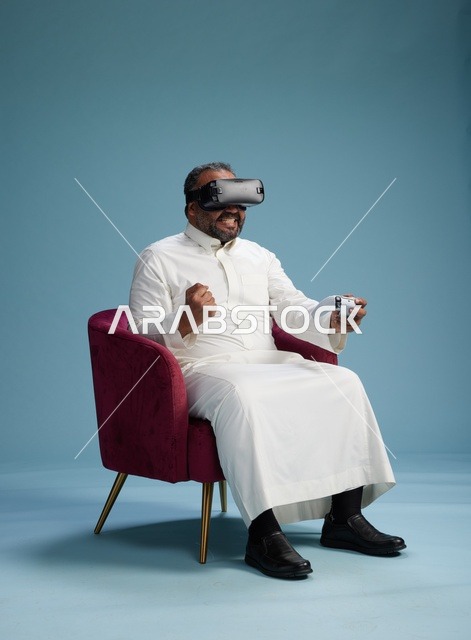 A portrait of a Saudi man wearing virtual reality glasses and holding a controller, enjoying video games, with a blue background.