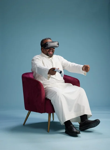 A portrait of a Saudi man wearing virtual reality glasses and holding a controller, enjoying video games, with a blue background.