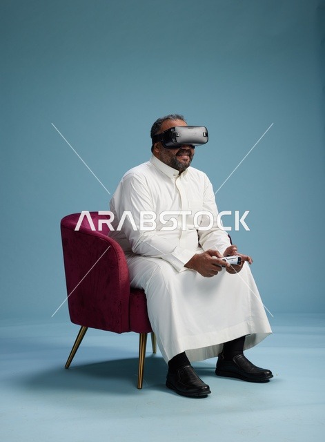 A portrait of a Saudi man wearing virtual reality glasses and holding a controller, enjoying video games, with a blue background.