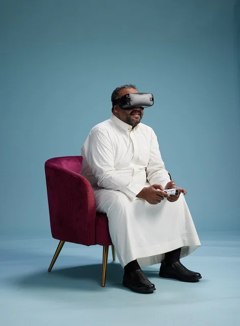 A portrait of a Saudi man wearing virtual reality glasses and holding a controller, enjoying video games, with a blue background.