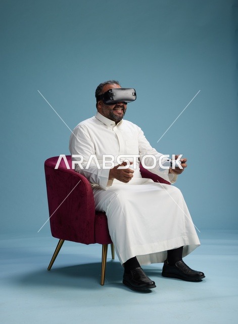 بورتريه لرجل سعودي يرتدي نظارة واقع افتراضي ويمسك بوحدة تحكم, الاستمتاع بالعاب الفيديو, خلفيه زرقاء