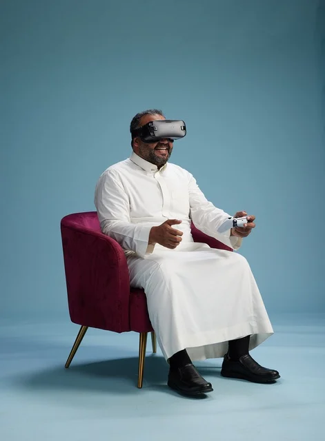 A portrait of a Saudi man wearing virtual reality glasses and holding a controller, enjoying video games, with a blue background.