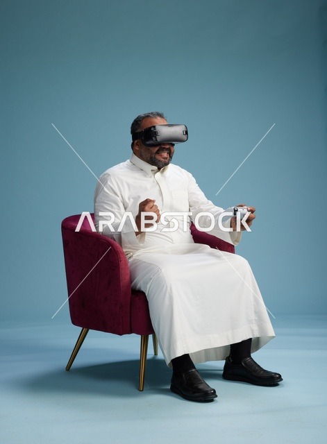 A portrait of a Saudi man wearing virtual reality glasses and holding a controller, enjoying video games, with a blue background.