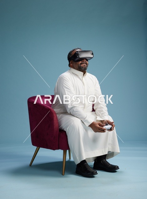 A portrait of a Saudi man wearing virtual reality glasses and holding a controller, enjoying video games, with a blue background.