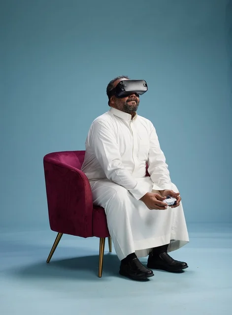 A portrait of a Saudi man wearing virtual reality glasses and holding a controller, enjoying video games, with a blue background.