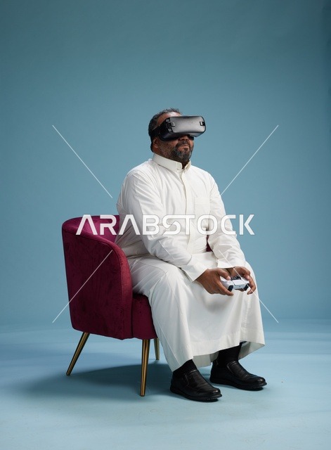 A portrait of a Saudi man wearing virtual reality glasses and holding a controller, enjoying video games, with a blue background.