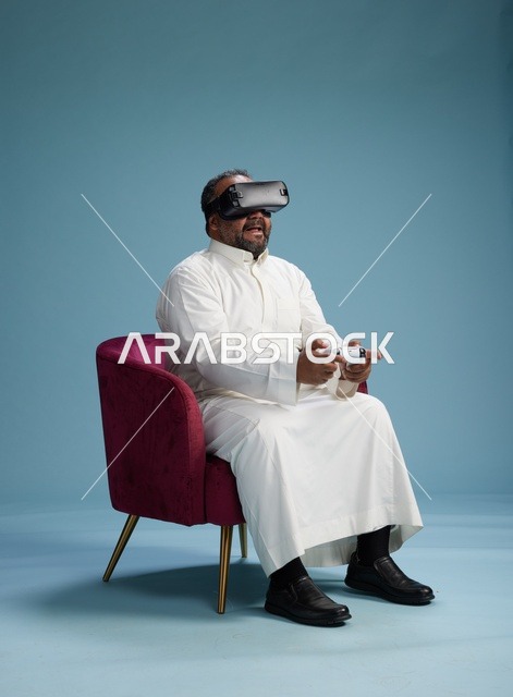 بورتريه لرجل سعودي يرتدي نظارة واقع افتراضي ويمسك بوحدة تحكم, الاستمتاع بالعاب الفيديو, خلفيه زرقاء