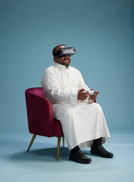 بورتريه لرجل سعودي يرتدي نظارة واقع افتراضي ويمسك بوحدة تحكم, الاستمتاع بالعاب الفيديو, خلفيه زرقاء