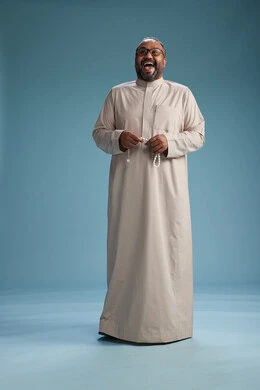 A portrait of a smiling Arab Saudi Gulf man wearing a beige thobe, holding a rosary in his hand and looking at the camera, full body, with a blue background.
