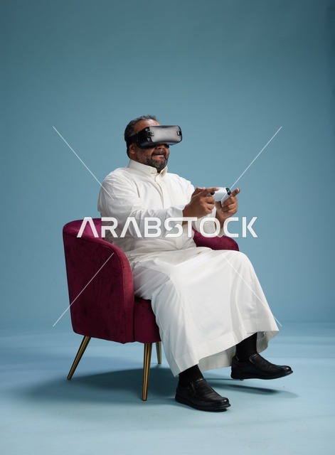 بورتريه لرجل سعودي يرتدي نظارة واقع افتراضي ويمسك بوحدة تحكم, الاستمتاع بالعاب الفيديو, خلفيه زرقاء