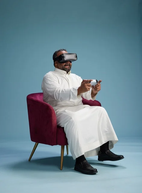 بورتريه لرجل سعودي يرتدي نظارة واقع افتراضي ويمسك بوحدة تحكم, الاستمتاع بالعاب الفيديو, خلفيه زرقاء