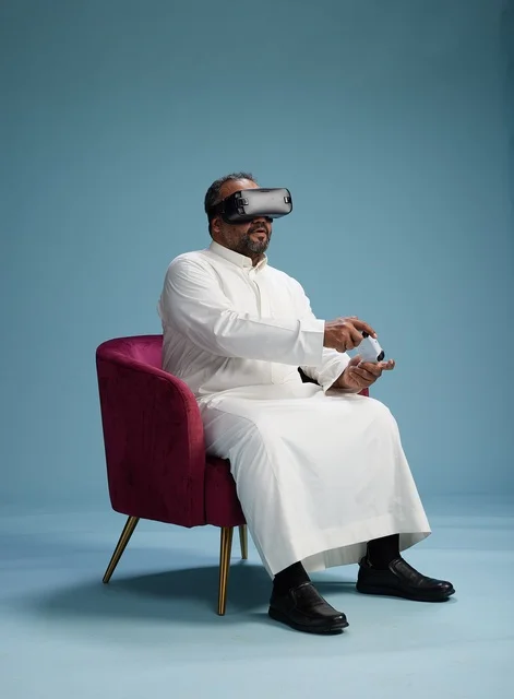 A portrait of a Saudi man wearing virtual reality glasses and holding a controller, enjoying video games, with a blue background.
