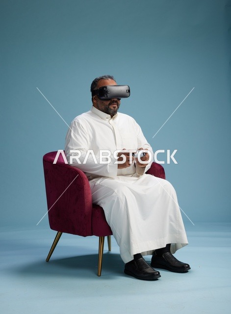 بورتريه لرجل سعودي يرتدي نظارة واقع افتراضي ويمسك بوحدة تحكم, الاستمتاع بالعاب الفيديو, خلفيه زرقاء